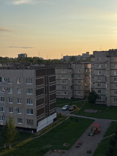 The image captures a twilight view of a residential area in Kirishi, Russia. The sky is a soft gradient of pale blue and warm orange, indicating the setting sun. In the foreground, two multi-story apartment buildings, constructed with grey brick and featuring numerous windows and balconies, dominate the scene. The building on the left has a distinct geometric pattern of dark brown panels on its facade. In front of the buildings, there is a patch of green grass, a paved walkway, and a small children's play area with a red and yellow playhouse. Two cars, a silver sedan and a dark SUV, are parked on the asphalt. A lone figure in light-colored clothing is walking on the grass pathway. In the background, more apartment buildings and trees are visible, with utility poles and distant structures also discernible. The overall impression is a quiet, ordinary evening in a Soviet-era housing complex.
