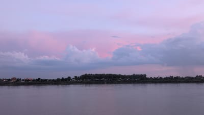 The image depicts a serene sunset over a wide, calm river with a village nestled along its opposite bank. The sky is painted with soft hues of purple and pink, interspersed with wispy clouds that catch the fading sunlight. A line of trees and structures, including buildings and what appears to be a water tower, form the backdrop of the village. The river's surface reflects the muted colors of the sky, creating a peaceful and tranquil atmosphere. This scene is characteristic of the Mekong River in Vientiane Capital, Laos, where sunsets often cast a similar magical glow over the landscape.