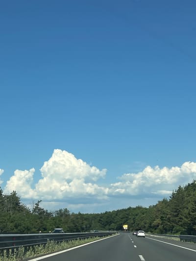 The image depicts a highway scene on a clear, sunny day. The sky is a vibrant blue, with fluffy white clouds scattered across it. The highway itself is asphalt, with white lane markings. On the right side of the road, there is a dense line of green trees. A metal guardrail separates the highway from the trees.

Several vehicles are visible on the highway. In the distance, a white car is traveling in the same direction as the viewer. Closer to the foreground, a yellow vehicle with flashing lights is stopped on the side of the road, possibly indicating some kind of road work or emergency. Another dark-colored vehicle is also present, partially obscured by trees.

The overall impression is one of travel and a bright, pleasant day. The lush green trees suggest a rural or semi-rural setting. The presence of the yellow vehicle with flashing lights suggests a potential disruption or event occurring on the highway. The time of day appears to be midday, given the bright sunlight and the clear sky. There is no visible text in the image.