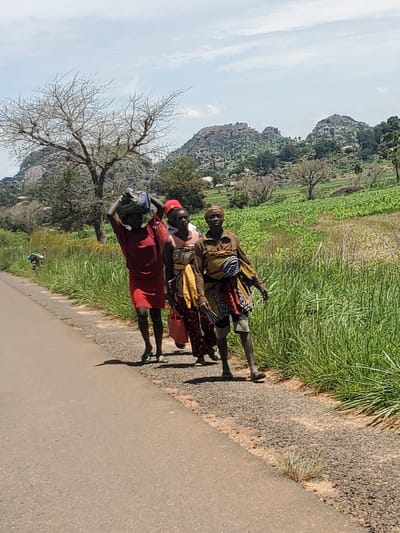 The image depicts four women walking along a paved road, carrying bundles on their heads. The road is bordered by a gravel shoulder and lush green vegetation. In the background, a hilly landscape with rocky outcrops and scattered trees is visible, along with agricultural fields. The sky is partly cloudy. The overall scene suggests a rural or semi-rural environment, possibly in the vicinity of Pankshin, Nigeria, given the geographical context. The women are dressed in colorful clothing, and their posture indicates they are engaged in travel or transport.