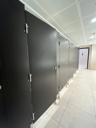 This image depicts the interior of a clean, modern men's public restroom located in Dorno, Italy. The photograph is taken from a low-angle perspective, looking along a lengthy row of black toilet stall doors that recede into the background. Each stall door is equipped with silver-colored hinges and visible latches or locks.
On the left side of the frame, a wall covered in white square tiles extends parallel to the stalls. To the right, a contrasting wall with light beige or grey square tiles is visible, from which white cylindrical supports extend, anchoring the stall partitions. The ceiling is white with a grid pattern, and some light fixtures or vents are discernable.
In the background, a doorway with a clear or frosted glass panel is visible, displaying a prominent male restroom symbol. The restroom appears empty, with no individuals present or any specific activity occurring. The space is brightly lit, and the surfaces of the black stall doors show subtle reflections. The overall impression is one of cleanliness, functionality, and a contemporary design aesthetic.