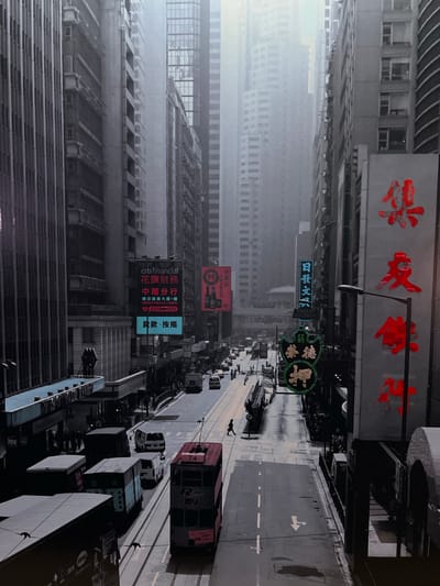 The image depicts a bustling city street scene, not Schoonderbuken, Belgium, but rather what appears to be a dense urban environment with tall buildings and significant traffic. The perspective is from a high vantage point, looking down a street lined with commercial buildings.

In the foreground, a red and grey double-decker tram is visible, numbered 74, moving down the street. Other vehicles, including vans and what appear to be trucks, are also present on the wet street, suggesting recent rain or damp conditions. Pedestrians are scattered along the sidewalks and crossing the street.

The buildings are characterized by their verticality and numerous windows, with a mix of architectural styles. Several large signs with Chinese characters are prominent on the buildings, indicating a likely location in East Asia, not Europe. Some signs are illuminated, adding to the vibrant, urban feel of the scene. The sky appears hazy or overcast, contributing to a somewhat muted color palette, though the red signage stands out distinctly. The overall impression is of a dynamic and active urban environment.