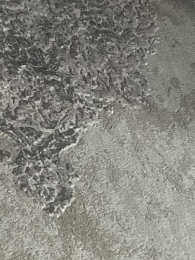 The image is a close-up, high-angle shot of a textured surface, likely pavement or rough stone, in a gray, monochromatic tone. The texture is irregular, with variations in light and shadow creating a mottled appearance. The texture is denser and darker on the left side of the image, appearing to recede towards the right, where it becomes more uniform and lighter. No people, subjects, objects, actions, or interactions are discernible in the image. The scene is devoid of any recognizable landmarks or features that would indicate a specific location within Rosenovo, Bulgaria, or any city.
