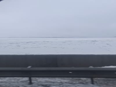 The image depicts a winter landscape in Rumbenieši, Latvia, characterized by a muted color palette under an overcast sky. In the immediate foreground, a dark gray metal guardrail is positioned horizontally, with some snow accumulation on the ground beneath it. Behind the guardrail, a dark gray, solid barrier, appearing to be concrete or a similar material, runs across the mid-bottom section of the frame. Beyond this barrier, a large body of water is visible, covered extensively in ice and snow, displaying an irregular surface texture consistent with partially frozen or broken ice. On the distant horizon, a low, dark landmass is faintly discernible, with a slender, dark vertical structure present on the far left edge of this distant shoreline. The sky is a uniform light gray, blending into the horizon without distinct cloud formations. No individuals or active interactions are present in the scene.