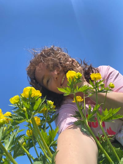 A low-angle, slightly upside-down shot shows a person with curly brown hair and a pink t-shirt holding yellow globe flower stalks against a clear blue sky. The person's face, with a visible smile, is partially framed by the yellow flowers and green foliage. The location is not discernible from the image.