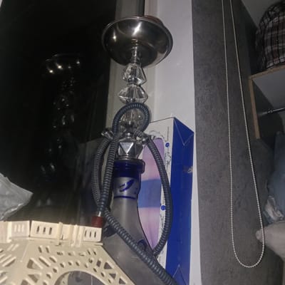 The image shows a hookah with a clear glass base and a metal stem with clear glass embellishments. Two black hoses are attached to the hookah. A blue and white cardboard box is partially visible behind the hookah. In the foreground, a portion of a white, decorative Eiffel Tower replica is visible. The background features a textured grey wall, a white wall, and a glimpse of a plaid garment on a shelf. The scene appears to be indoors, in what could be a domestic or retail setting. The location is identified as Бәйтерек, Kazakhstan.