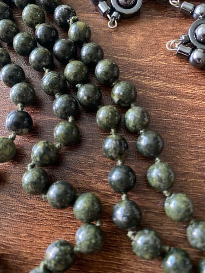A close-up, overhead shot shows several strands of beaded jewelry on a wooden surface. The beads are mostly round and appear to be made of a dark green, mottled stone, possibly jade or serpentine. The beads are threaded onto a light-colored string, with visible knots between each bead. Some of the beads have a glossy sheen, reflecting the light. In the upper right portion of the image, there are two dark, metallic clasps or connectors, each with smaller metallic rings attached, suggesting the jewelry is a necklace or bracelet. The wooden surface has a visible grain pattern and a warm, brown hue. The lighting is direct, creating highlights and shadows on the beads and the wood.