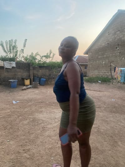 The scene depicts an outdoor residential area in Idiroko, Nigeria, during either sunrise or sunset, indicated by the soft, warm light in the sky.
A single adult female is positioned centrally in the foreground, facing the camera with her head turned. She has short, dark hair and is dressed in a dark blue spaghetti strap top and an olive-green mini-skirt. A light-colored flip-flop is visible on her left foot. She holds a smartphone with a red or pink case and a blue screen in her right hand.
To the left and behind the female subject, a low concrete block wall is present, with white fabric draped over its top. At the base of this wall, on the dirt ground, are several containers: two blue plastic barrels with lids, one yellow bucket, and one grey bucket. Sparse green vegetation, including small trees, is visible beyond the wall.
To the right of the subject, a building constructed of unplastered concrete blocks is partially visible, featuring a sloped roof made of reddish-brown corrugated metal. A clothesline extends from this building, suspending multiple items of multi-colored clothing for drying. In the background, another building with a reddish-brown corrugated metal roof is partially observable. The ground throughout the visible area is unpaved dirt with minor scattered debris.