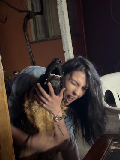 The image shows a woman and a dog in what appears to be an outdoor setting at night. The woman, with long dark hair, is holding the dog close, with her eyes closed and mouth open in an exaggerated expression. The dog, a German Shepherd, has its mouth open as well, revealing its teeth. Both the woman and the dog are close to the camera, making them the central focus. The background is dark and somewhat blurry, but suggests an urban environment with buildings and some vegetation. The presence of a wooden table in the foreground and a white chair in the background hints at a patio or outdoor seating area. There is a vine-like structure with dark branches visible on the left side of the image. The overall lighting suggests artificial light sources, possibly from nearby buildings or lamps. The location of Golyam Chardak, Bulgaria is not evident from the visual cues in the image itself.