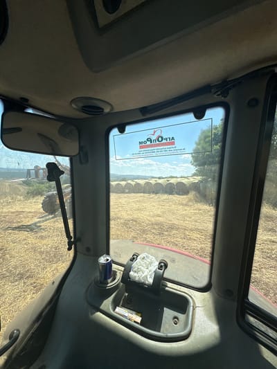 The image shows the interior view from the driver's cabin of a tractor. The cabin is dusty and shows signs of use. A can of Red Bull is placed in a cup holder, next to a crumpled white cloth. Outside the window, a field is visible, covered with hay. Several round bales of hay are lined up in the distance. A large tractor is also visible in the field to the left, and a piece of agricultural machinery is attached to it. The sky is blue with some clouds. A sticker with the logo "Moprorm" is affixed to the windshield. The overall impression is of a farm setting on a sunny day.