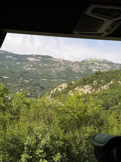 The image shows a view from a gondola lift cabin in Markovići, Montenegro. A steep, rocky mountain covered with green vegetation dominates the scene. Cable support towers and gondola cars are visible against the mountain. The sky is partly cloudy. The interior of the gondola car frame is visible on the left and right of the image.
