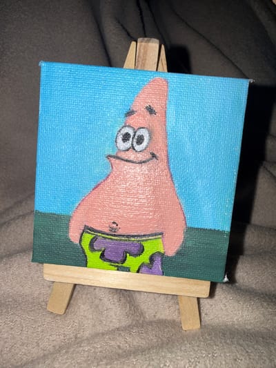 A small, square canvas painting featuring Patrick Star from SpongeBob SquarePants is prominently displayed on a miniature wooden easel. Patrick is depicted with a pink body, large white eyes with black pupils, a black smiling mouth, and a small black belly button. He wears green shorts with purple, abstract patterns. The painting's background consists of a bright blue upper section, representing the sky, and a dark green lower section, representing the ground. The canvas exhibits a visible woven texture. The easel, constructed from light-toned wood, rests on a soft, textured surface that appears to be a brownish-grey blanket or carpet. Shadows are cast to the right of the easel and painting, suggesting an overhead or left-side light source. No people, discernible actions, or interactions are present in the scene. The image provides no contextual information to determine its geographical location in the city of Dhadna, United Arab Emirates.