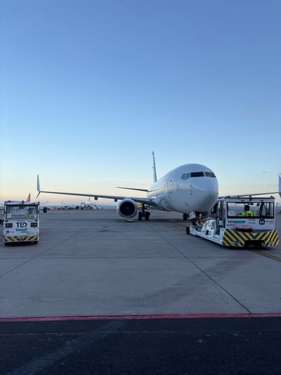 The image depicts a commercial aircraft, specifically a Boeing 737, at Tenerife South Airport (TFS) in Granadilla de Abona, Spain. The aircraft is parked at a gate and connected to a pushback tractor. There is a boarding bridge. The sky is visible with a clear blue and orange tint, suggesting either sunrise or sunset. The concrete apron surface shows some staining and wear. A worker is visible inside the pushback tractor.