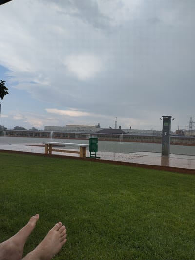 This image depicts an outdoor scene, likely in a park or recreational area, with a body of water and a grassy area in the foreground. The sky is overcast with gray clouds, and it appears to be raining, with visible raindrops on the surfaces. In the foreground, the bare feet of a person are visible lying on lush green grass. Behind the grass, a wooden deck or walkway is visible, with a green trash bin and a wooden bench. Beyond this, a large body of water is present, featuring a fountain. Along the edge of the water, a covered structure with seating is visible. In the background, industrial buildings, smokestacks, and electrical towers can be seen, suggesting proximity to an industrial zone. The location is identified as Romanivka, Ukraine.