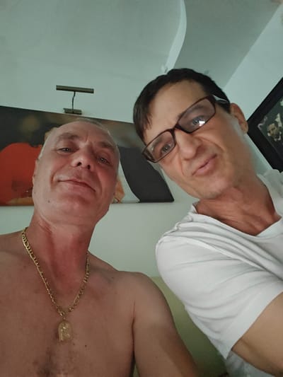 The image is a selfie featuring two men, likely friends or acquaintances, in an indoor setting. The man in the foreground on the left is facing the camera directly, with a slight smile. He is shirtless and wearing a thick gold chain with a pendant. His skin appears slightly reddened, possibly from sun exposure. The man on the right is also looking towards the camera, wearing glasses and a white t-shirt. He has a more dynamic expression, with his mouth slightly open and eyes looking intently at the viewer.
Behind the men, the background is somewhat blurred but suggests an interior space. There is a piece of abstract art on the wall featuring a large orange shape, and a framed picture of Abraham Lincoln is visible to the right of the man with glasses. A simple lamp is mounted on the wall above the man in the foreground. The lighting suggests it could be daytime, but no definitive clues about the time of day or weather are present. The overall mood seems relaxed and informal, characteristic of a personal photograph. There is no visible text in the image.