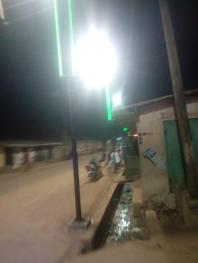 The image depicts a street scene at night in Tundun Wada West, Nigeria. A tall, lit signpost with bright green vertical lights stands prominently on the left side of the road, illuminating the surrounding area with an intense glow. The road itself appears to be unpaved and slightly damp, with a visible drainage channel running alongside it.

To the right of the road, a building with a light-colored wall is visible. On this wall, there are several markings, including what appears to be a graphic of a mobile phone or a card reader. Above the graphic, there are three blue circles and the word "first" written in yellow. A green door is also visible on this building.

Further down the street, on the left side, blurred figures and a motorcycle are discernible, suggesting some activity and movement. The overall atmosphere is dark, characteristic of nighttime, with the bright artificial lights providing the primary illumination. The scene suggests a commercial or residential area in a developing locality, with the street serving as a central thoroughfare. No specific weather conditions are evident, but the dampness of the road might imply recent rain. The image conveys a sense of everyday life occurring at night in this Nigerian location.