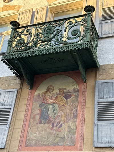 The image captures a building in Riedlingen, Germany. A decorative, green wrought-iron awning with ornate embellishments extends over a window. To the right of the awning, on the wall, is a faded painting depicting a religious scene, possibly the Flight into Egypt, with figures including a woman, a child, and a man on horseback. Below the painting is a light grey, shuttered window. Another partially visible shuttered window is above the painting. The building has a light yellow facade with variations in texture and color.