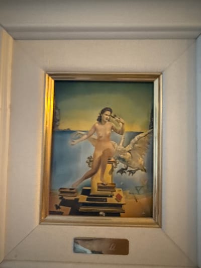 The image shows a framed surrealist painting featuring a nude woman and a large bird against a dreamlike landscape. The woman, with dark hair and a fair complexion, is positioned on a stack of books or platforms. She is holding a bird or a similar object in her right hand and her left arm is extended forward. To her right, a large, feathered bird with its wings spread appears to be flying or swooping down. The background depicts a body of water, a distant coastline with structures, and a gradient sky transitioning from blue to yellow. The painting is enclosed in a golden frame, which is then placed within a larger, white matting and frame. A small, rectangular plaque is affixed to the bottom of the white matting, featuring some indistinct lettering. The overall presentation suggests the artwork is displayed on a wall, likely in a domestic or gallery setting. The image does not contain any information that would allow for identification of its location in the city of Yukki, Russia.