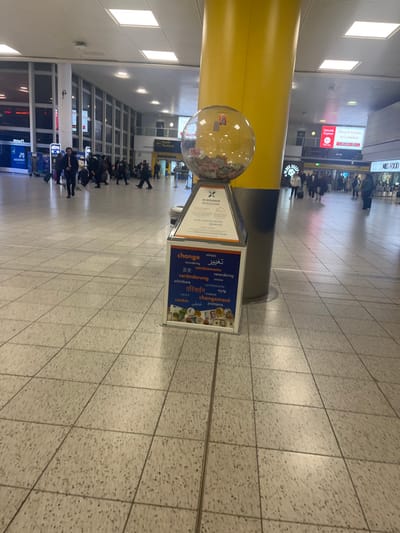 This image captures a bustling indoor scene, likely an airport terminal given the context of Horley, United Kingdom, which is home to Gatwick Airport. The setting features a wide, tiled floor and a high ceiling with bright rectangular lights. A large, bright yellow cylindrical pillar dominates the left side of the frame.
The main subjects are numerous people, mostly seen in the background and walking across the floor. They appear to be travelers, some with luggage, moving through the terminal.
In the foreground, to the right of the yellow pillar, is a prominent stand. It features a transparent spherical container filled with banknotes and coins, possibly a collection point for donations or unwanted foreign currency. Below this sphere is a display board. One side of the board shows text in various languages for "change," including English, Arabic ("تغییر"), German ("veränderung"), Romanian ("schimbare"), Dutch ("verandering"), Hindi ("परिवर्तन"), French ("changement"), Spanish/Italian ("cambio"), Urdu ("تبدیلی"), and Serbo-Croatian/Bosnian/Croatian ("promjena"). The other side of the display has a logo with an 'X' and text that includes "YOUR LONDON ARRIVAL" and "Continental," further indicating an airport environment.
Other visible text on overhead signs includes "M&S FOOD" and "Direct trains to London," clearly indicating transport links from this location. Another sign reads "North Terminal," specifying a particular section of the airport. The word "atm" is also partially visible on a sign. The time of day cannot be determined as the scene is entirely indoors under artificial lighting. The overall atmosphere appears busy and functional.