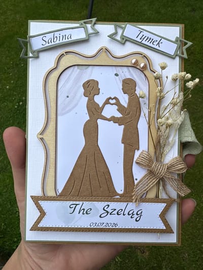 A handmade wedding card is presented, held by a person's fingers against a background of green grass. The card features a central cutout of a bride and groom forming a heart shape with their hands, rendered in a brown silhouette. Above the couple, two banners display the names "Sabina" and "Tymek" respectively, in a stylized font. Below the couple, another banner reads "The Szeląg" with a date "03.07.2026" underneath. The card is embellished with small dried flowers, a burlap bow, and subtle pearl accents. The card is positioned vertically, with the top edge slightly tilted away from the viewer. There is no discernible connection to Belfast City, United Kingdom.
