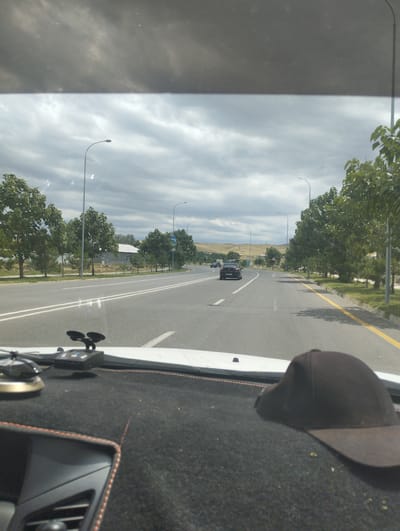 The image is a first-person perspective from inside a vehicle, looking out through the windshield. The vehicle is traveling on a multi-lane paved road lined with trees and streetlights. In the distance, there are some buildings and rolling hills under a cloudy sky. The dashboard of the car is visible in the foreground, with a black baseball cap and a black electronic device with suction cups. The location is identified as Birlashgan, Uzbekistan.