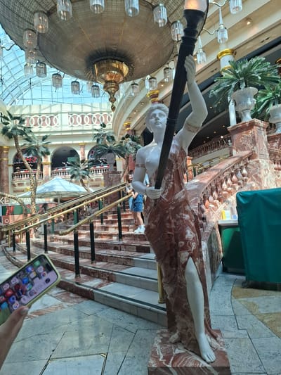 A full shot shows a large marble statue of a woman holding a torch, located in the Trafford Centre in Manchester, United Kingdom. The statue is positioned on a marble staircase with a red and white marbled pattern. Behind the statue, palm trees and ornate railings of the upper levels of the shopping mall are visible. The ceiling features a large, decorative chandelier and a glass roof. A person's hand holding a smartphone is in the foreground, obscuring the bottom left corner of the image. Two young women are walking up the stairs to the right of the statue. The environment appears to be an indoor shopping mall with a grand architectural design.