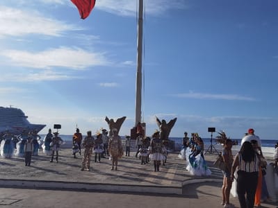 The image shows a group of people performing a traditional dance in front of a flagpole in the city of Cozumel, Mexico. The location is approximately the Parque Benito Juárez on the waterfront, based on the coordinates provided. The performers are wearing colorful costumes and are moving in a synchronized manner. There are several spectators watching the performance. A cruise ship is visible in the background. The scene is taking place on a paved outdoor stage. The sky is clear and blue, with some clouds in the distance. There are two bronze statues of birds in the background. The flagpole is flying a Mexican flag that is not fully extended. The scene is lively and festive, with a sense of celebration and community.