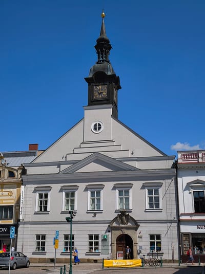 The image shows a street scene in Vysoké Mýto, Czechia, dominated by a grand building with a prominent clock tower. The building, likely a town hall or a similar civic structure, features a light grey facade with arched doorways and multiple windows. The clock tower, topped with a spire and a golden finial, has a dark, ornate clock face with Roman numerals. Below the clock, architectural details include a round window and stepped rooflines.
In front of the building, a paved sidewalk is visible, with parked bicycles and a yellow banner displaying the text "(M)ýto žije!". Several people are present: a person in a blue dress walks across the street carrying a package, another person stands near an open doorway, and a few individuals are seen through the windows of the building. A gray car is parked on the left side of the street, next to a sign with a "P" symbol. Adjacent buildings, one with decorative yellow and gold elements and another with a white facade and red railings, frame the central structure. The sky is a clear, bright blue, suggesting a sunny day, likely mid-day given the strong shadows. The overall impression is of a vibrant historical town center.