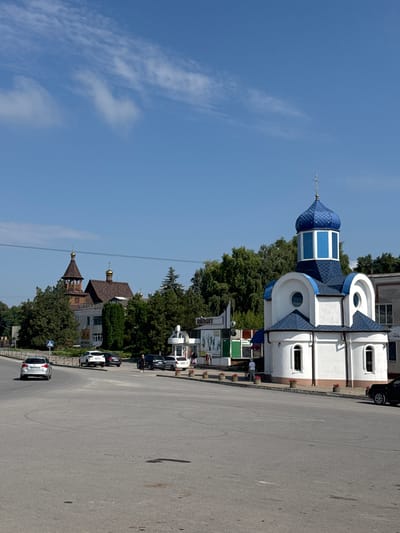 A photograph taken on a sunny day in Chemerivtsi, Ukraine, depicts a street scene with two churches and several cars. In the foreground, a paved road with tire marks curves to the right. To the right of the road is a small, white church with a blue, multi-faceted dome topped by a cross. The church has arched windows. To the left of this church, further down the street and partially obscured by trees, is a larger, older-looking church with a brown, multi-tiered bell tower topped by a golden dome and a smaller, brown-domed structure. Several cars are parked or driving on the road. One silver car is in the far left lane, and a white car is parked on the right shoulder. In the middle ground, there are trees and several buildings, including what appears to be a kiosk or small shop with colorful panels. A pedestrian is visible walking away from the white church. The sky above is a clear blue with a few wispy white clouds.