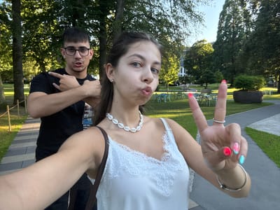 In a park in Bad Orb, Germany, a young woman is taking a selfie outdoors. She is wearing a white lace-trimmed camisole and a shell necklace. Her hair is dark brown, and she has bright pink and turquoise fingernails, with one hand raised in a peace sign. A young man with dark hair and glasses is standing behind her, wearing a black t-shirt. He is gesturing with his left hand, with his index finger and thumb forming a "rock on" sign. The background shows trees, grass, and a paved path.