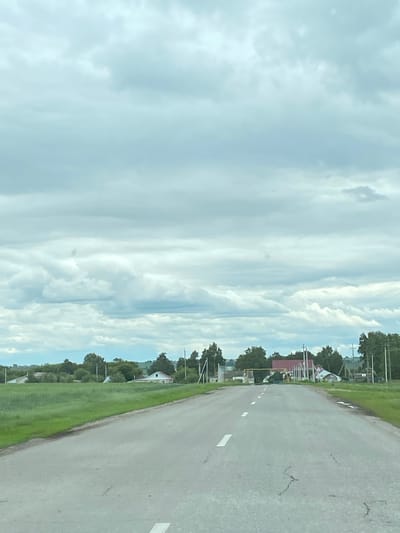 An asphalt road stretches into the distance through a rural landscape under a cloudy sky. The road has white dashed lines marking the lanes. On either side of the road is a vibrant green field. In the middle ground, a cluster of buildings is visible, including houses with red roofs, and trees. The sky is filled with gray and white clouds, with some brighter patches suggesting sunlight breaking through. The scene appears to be in or near the village of Nadezhdino, Russia, as indicated by the environment and architectural style. There are no people visible in the image.