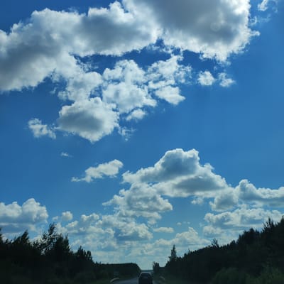 The image depicts a view from a moving vehicle on a road lined with dark green trees on either side, suggesting a rural or forested area. The sky is a vibrant blue, filled with numerous fluffy white clouds of varying sizes, some casting shadows on themselves. The sun is visible in the upper right corner, partially obscured by a large cloud, creating a bright spot in the sky. In the foreground, the road stretches ahead, with a dark-colored car visible in the distance. The overall scene conveys a sense of travel on a clear day. The provided metadata indicates this scene is located in Kineshma, Russia.