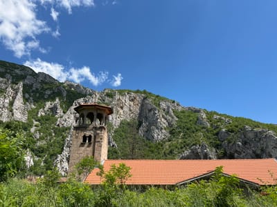 The image depicts a bell tower and a red-tiled roof of a building, set against a backdrop of a rocky, tree-covered mountainside under a clear blue sky with scattered clouds. The bell tower is made of stone and features arched openings and a decorative top. Lush green vegetation, including tall grass and bushes, occupies the foreground, partially obscuring the lower portions of the building. The scene is static and appears to be a landscape shot. The location is identified as the city of Lyutibrod, Bulgaria.