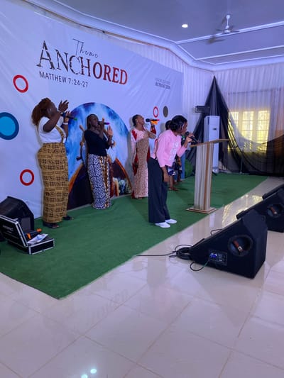 **Scene:** A well-lit interior space, likely a church or event hall in Miango, Nigeria. The backdrop prominently features the word "ANCHORED," followed by "MATTHEW 7:24-27" and "YOUNG COUPLES RETREAT 2026." A nautical theme is suggested with a ship's wheel graphic.

**People:** Four women and one child are standing on a green carpeted stage, facing toward the right of the image. Each holds a microphone. From left to right:
*   A woman with dark hair wearing a white top and a brown and yellow patterned skirt.
*   A woman with short hair wearing a black top and a black and white patterned skirt.
*   A woman with long hair wearing a red and white patterned dress.
*   A woman with short hair wearing a pink sweatshirt and dark pants.
*   A child standing near the podium wearing a blue shirt and jeans.

**Objects:**
*   A wooden podium is located at the right of the stage.
*   Two large black speakers stand to the right of the stage.
*   An equipment case is on the floor to the left.
*   White curtains drape the ceiling.

**Actions:**
*   The women are standing at the front of the stage holding microphones.

**Environment:**
*   The floor is tiled.
*   The space appears to be indoors.
*   Good lighting.