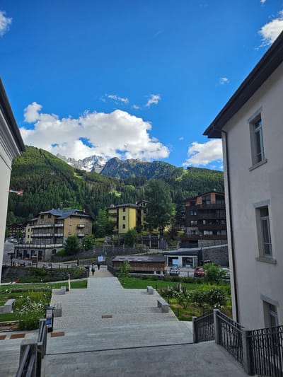 This vertical image, framed by the eaves and walls of a building, captures a vibrant, sunlit mountain village scene, likely in Ponte di Legno, Italy. The vantage point is slightly elevated, looking down into a valley.

In the background, majestic mountains dominate, their peaks partially covered with snow, indicative of a high-altitude or early season setting. The lower slopes of the mountains are densely forested with dark green coniferous trees. The sky above is a brilliant blue, dotted with scattered, fluffy white cumulus clouds. A faint white contrail from an aircraft is visible in the upper left quadrant of the sky.

The midground and foreground are occupied by various multi-story buildings, showcasing a blend of traditional and modern alpine architecture. Some structures feature light-colored plaster facades with numerous windows and balconies, often adorned with metal railings. Other buildings exhibit contemporary designs with dark wooden cladding. Between these structures, well-maintained green lawns, landscaped flowerbeds with white and purple blossoms, and scattered trees add to the picturesque setting.

A wide, paved square or walkway, constructed from light grey stone tiles and cobblestones, stretches across the right side of the foreground, featuring stone benches and streetlights. Several cars, including a distinct red vehicle, are visible on a road or parking area in the midground. Two distant figures appear to be walking on a paved pathway. A blue signpost with white symbols, including a 'P' for parking, a wheelchair accessibility symbol, and partially discernible text (possibly "BUS"), stands near the paved area.

The clear sky, bright sunlight, and lush greenery suggest a pleasant day, likely in late spring or summer. The overall impression is one of a serene and well-developed mountain resort town.