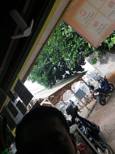 The image shows a close-up view of a person's head and shoulders, likely a man, with a black motorcycle helmet and a motorcycle in the foreground. The person is standing in front of a shop in Kecamatan Babakan Madang, Indonesia, with coordinates -6.561 latitude and 106.863 longitude. The shop is a small, open-air structure with a sign advertising various services, including "Pulsa & Paket Data" and "Top Up ShopeePay". The shop is located under a large tree with green leaves, which provides shade for the area. There are several people in the background, including a group of children standing near a motorcycle, and the shop has a wooden roof and a white wall. The environment is a suburban area with a paved road. The person is likely waiting for someone or something, as the scene is seemingly mundane. The image was taken from a low angle, likely by someone sitting on the motorcycle.