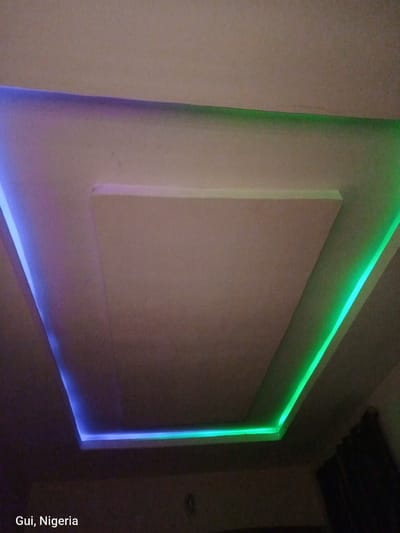 A ceiling with recessed lighting features is shown in a dimly lit room. The ceiling is painted a neutral color and has a central rectangular depression. LED lights are installed along the edges of the recessed area, casting a gradient of blue, purple, and green light. The lighting creates a modern and atmospheric effect. In the bottom left corner of the image, the text "Gui, Nigeria" is visible. The overall impression is of an interior space with contemporary design elements.