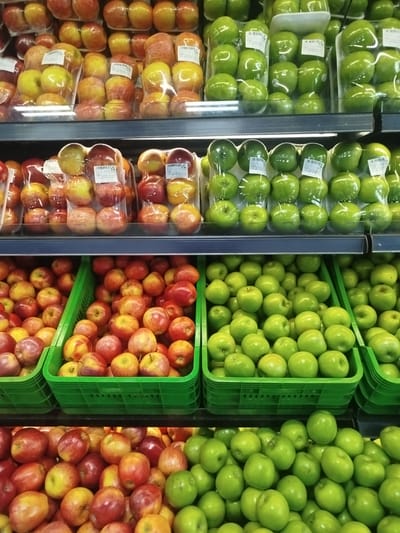 This media file captures a brightly lit supermarket produce section, showcasing a variety of apples displayed across multiple shelves. No people are visible in the image.

The scene is dominated by rows of apples, both pre-packaged and loose in green plastic crates. The top two shelves feature apples individually wrapped in clear plastic, likely in sets, with white labels. The topmost shelf on the left displays red and yellow apples, while the right side of the same shelf holds vibrant green apples. The shelf directly below it follows a similar pattern, with red and yellow apples on the left and green apples on the right, all uniformly packaged.

Below these pre-packaged rows, two lower shelves are filled with loose apples contained within numerous green plastic crates. The left side of these lower shelves predominantly features red and yellow apples, while the right side is entirely dedicated to piles of green apples. This arrangement creates a visual gradient of color across the display.

Notable details include the clear plastic packaging that encases groups of apples, presumably for sale by weight or count. Many of these packages bear white adhesive labels with black text. Partially visible text on these labels includes "PRECIO" (Price), "1 KG" (indicating 1 kilogram), and specific prices such as "Bs. 1.20" and "Bs. 1.10", where "Bs." likely refers to Venezuelan Bolívares, consistent with the Barquisimeto, Venezuela location context. Several labels also explicitly state "PROD. DE CHILE" (Product of Chile), indicating the origin of the fruit. The apples appear fresh and are neatly organized, suggesting a well-maintained grocery store environment. The lighting is artificial and consistent throughout, typical of an indoor retail setting.