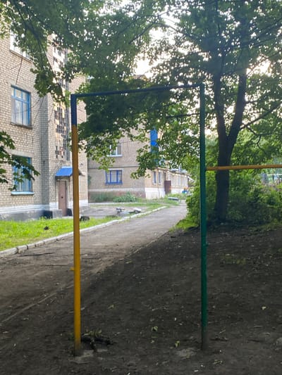 This image depicts an outdoor scene in the city of Antratsyt, Ukraine. In the foreground, a dirt patch is visible with fallen leaves and debris. Two vertical poles stand in the dirt: one is yellow and the other is green. A horizontal yellow bar is attached to the green pole. Behind the poles, a paved pathway runs diagonally across the image. To the left of the pathway, a grassy area with a curb separates it from a light-colored brick building. This building has multiple windows with blue frames and a blue-canopied entrance with a brown door. A large tree with lush green foliage is situated to the right of the pathway, partially obscuring another building in the background which also appears to be brick with blue-framed windows. The overall environment suggests a residential courtyard or park area.
