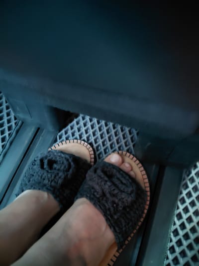 The image shows a close-up view of a person's feet wearing dark, crocheted slide sandals. The sandals have a tan sole with a decorative orange stitched edge. The feet are resting on a textured, diamond-patterned floor, likely inside a vehicle or on a platform. The background is dark and out of focus, suggesting an interior space. The image does not provide any visual cues or context that would indicate a specific location within the city of Молодёжный, Kazakhstan.