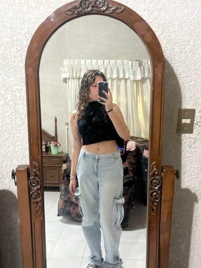 A person takes a mirror selfie. The mirror has a wooden frame with roses carved. She's wearing a black crop top and light-wash cargo jeans. There is a light switch above the mirror and beige curtains are in the background. A wooden dresser with a flower vase sits behind her.