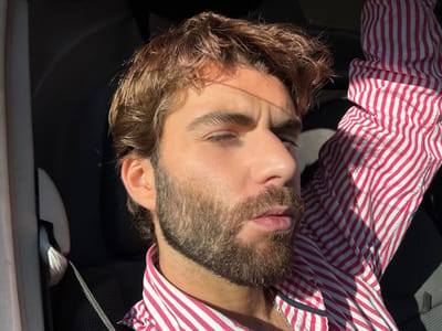 A male subject is depicted in a close-up portrait, located within the interior of a vehicle in Savigneux, France. The individual possesses brown, wavy hair and a full brown beard. He is wearing a long-sleeved shirt featuring vertical pink and white stripes, with a metallic chain visible at his neck. Strong natural light, consistent with direct sunlight, illuminates the right side of his face (viewer's left), causing a slight squint in his right eye. The left side of his face (viewer's right) and the vehicle's interior remain in shadow. A car seatbelt is visible across his left shoulder (viewer's right). The subject's right arm (viewer's left) is raised, with the striped sleeve prominently displayed. His head is slightly turned, and his lips are slightly parted.