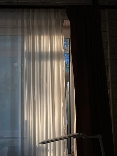 The image depicts a window covered by sheer white curtains on the left and a dark brown curtain on the right. Sunlight streams through the sheer curtains, creating strong vertical shadows and highlighting the texture of the fabric. A portion of a white desk lamp with articulated arms is visible in the foreground, positioned to the right of the center. Through the gap between the curtains, a window frame and a glimpse of the sky and some foliage are visible. The background wall appears to be made of light-colored bricks. The scene is set indoors, with the light suggesting it is daytime, possibly late afternoon given the warm tones of the sunlight. The specific location within Podilsk, Ukraine, cannot be determined from the visual information provided. There are no people or discernible actions taking place.