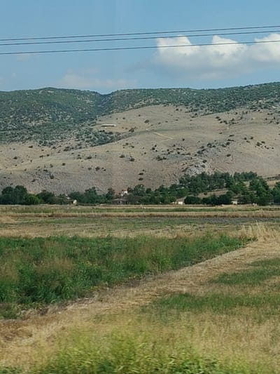 The image depicts a rural landscape with rolling hills in the background, covered in sparse vegetation and a dirt road winding up one of the slopes. A cluster of trees is visible in the mid-ground, with several small buildings nestled amongst them. In the foreground, there are fields of dry grass and some green vegetation. Power lines are visible in the sky. The overall scene suggests a sunny day in a semi-arid environment. The location is identified as Taxiarches, Greece. There are no people or animals visible in the image.
