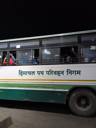 The image shows the side of a white and green Himachal Pradesh Road Transport Corporation bus at night, likely in Kajheri, India. The bus has tinted windows, and several passengers are visible inside, some appearing to be standing. Green text in Hindi, "हिमाचल पथ परिवहन निगम," meaning "Himachal Road Transport Corporation," is prominently displayed on the white section of the bus. Below the windows, there are compartments labeled "BATTERY BOX" and "SPARE WHEEL." A large tire and part of the wheel well are visible on the right side of the bus. The ground in front of the bus appears to be asphalt or concrete, with a raised concrete curb on the left. The overall lighting suggests an outdoor setting at night, possibly a bus stop or depot.