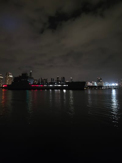 The image depicts the Coronado city skyline at night, reflected in calm water. The buildings are illuminated, casting light onto the water's surface. The sky is dark and partially obscured by clouds. There appear to be ships or other structures along the shoreline. The scene is quiet and still.