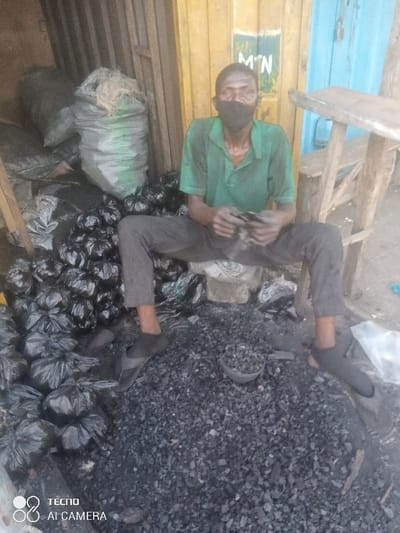 The image displays a charcoal vendor operating in Yalwa, Nigeria. An adult male, with dark skin and wearing a green short-sleeved polo shirt, dark trousers, and dark slippers, is seated on a low, possibly concrete, block. He wears a black fabric face mask covering his mouth and nose. His hands, soiled with charcoal dust, are positioned in front of him, appearing to manipulate small, dark objects. In front of the man, a large pile of loose, irregularly shaped charcoal pieces covers the ground. To his immediate left, a substantial stack of small, black plastic bags, each filled with charcoal, is visible. Behind him, two larger sacks, one grey and one light green, are partially visible, likely containing more charcoal or similar bulk material. A small, dark bowl or scoop containing charcoal pieces rests on the ground near his feet. The background features a weathered yellow metal wall or door, on which green lettering for "MTN" is partially visible. To the right, a section of a light blue door or shutter is present, alongside a crude, unfinished wooden table or stool. The ground throughout the scene is uneven, dusty, and scattered with charcoal fragments and debris.
