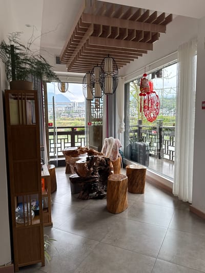 The image shows an interior scene, likely a living room, in Zhuoshui, China. The room features a decorative wooden ceiling, large windows offering a view of the town and distant mountains, log stools, and a large gnarled wooden table. A red papercut decoration hangs near the window. The floor is tiled. A wooden shelving unit holds books and decorative items. A potted plant is visible near the shelves. A suitcase is visible outside on the balcony. No people are visible.