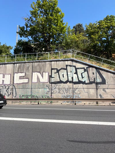 The image shows a concrete wall with graffiti on it, located in Bron, France. The wall is situated above a highway, with trees and a blue sky in the background. The graffiti consists of large, stylized letters. In the foreground, a section of the highway is visible, with a black car on the left side of the image. A metal guardrail runs along the top of the wall. The overall scene depicts an urban landscape with elements of street art.