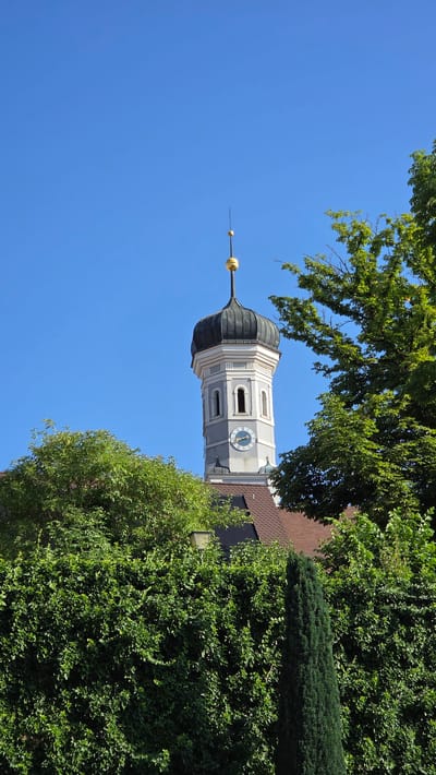 This is a photograph taken from a low angle, looking up at a church tower in Ulm, Germany. The tower is tall and slender, with a grey and white facade. It has a domed roof, topped with a golden spire. There are several arched windows on the tower, and a clock face is visible on one side. The tower is surrounded by lush green trees, with a clear blue sky in the background. The image appears to have been taken on a sunny day.